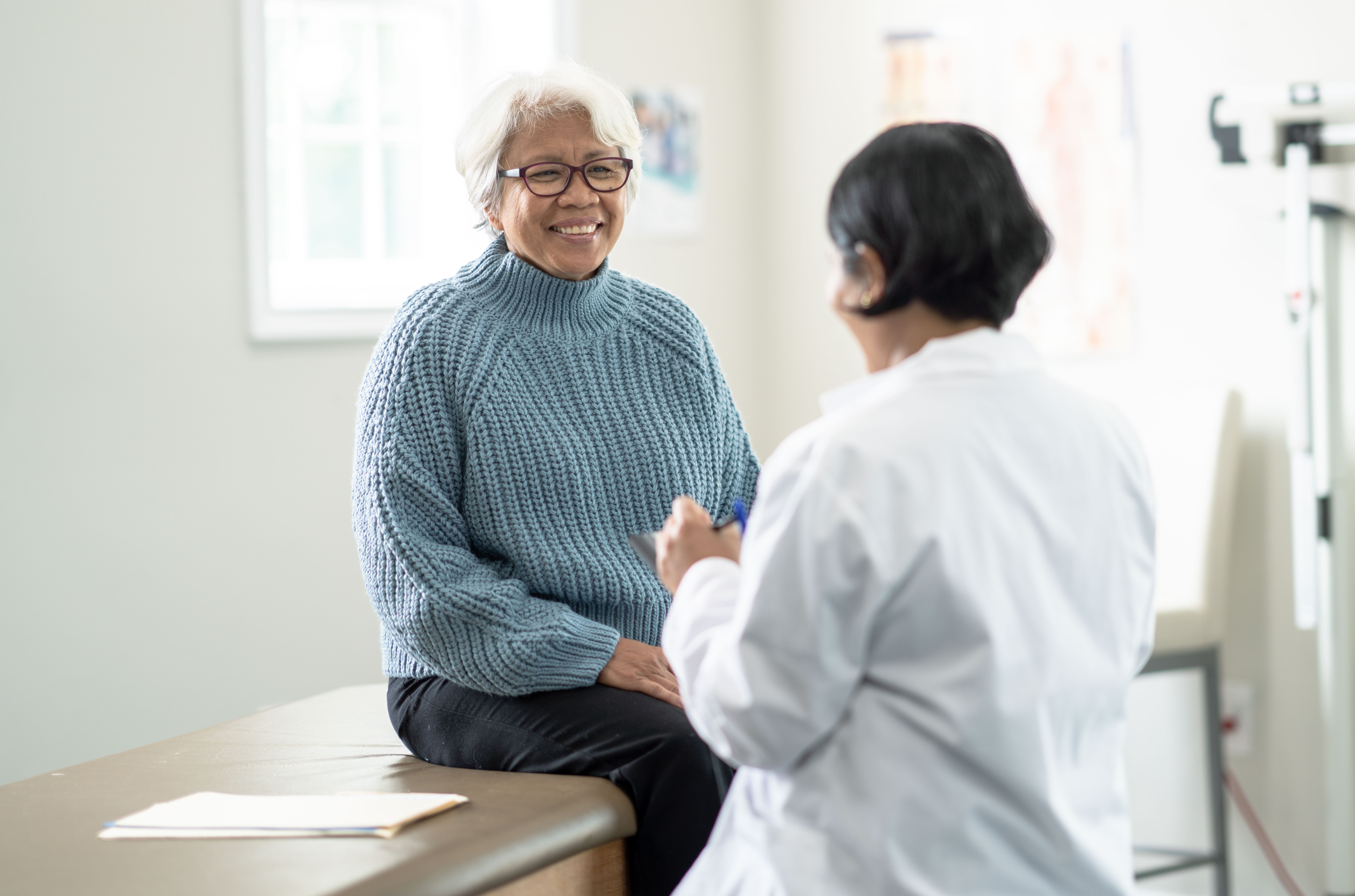 A senior woman sits up on the exam table at a doctors appointment.  She is dressed casually in a blue sweater and has a smile on her face as she glances at the doctor.  Her female doctor is wearing a white lab coat and seated in front of her as she takes notes on a tablet.